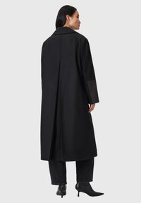 Trench coat nero con colletto, vestibilità ampia e maniche lunghe. Presenta una piega posteriore e dettagli neri a contrasto sulle maniche.