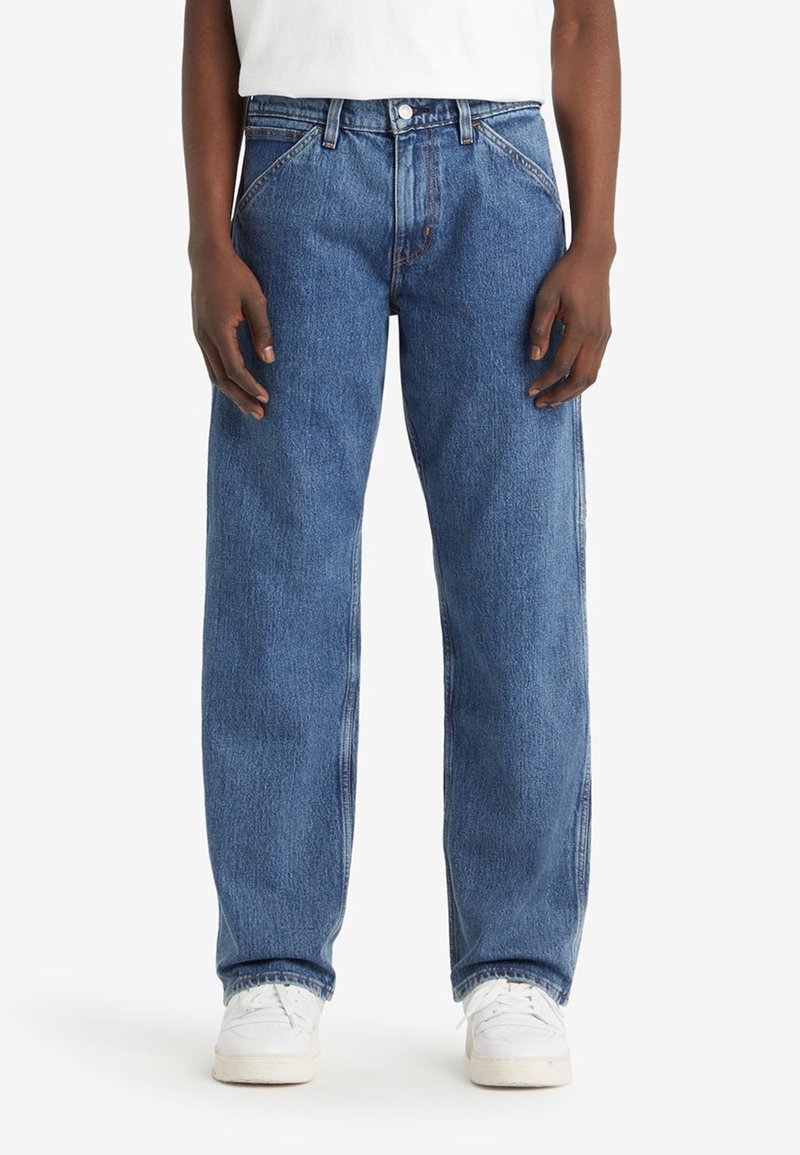 Personne portant un jean bleu ample et des baskets blanches, debout avec les mains détendues sur les côtés devant un fond uni.