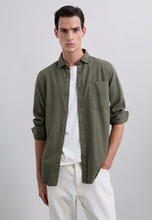 Camisa caqui de botones con una textura ligera, mangas enrolladas y un bolsillo en el pecho izquierdo, llevada sobre una camiseta blanca y combinada con pantalones claros.