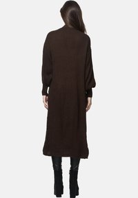 Lång, brun stickad cardigan med långa ärmar, med ett strukturerat mönster och öppen framsida. Stylen är komplett med svarta åtsittande byxor och stövlar.