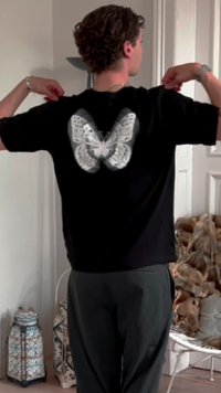 T-shirt nera con un grande grafico di una farfalla bianca sul retro. Il tessuto appare morbido e il design è centrato e dettagliato.