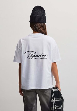 Person trägt ein weißes Pegador-T-Shirt und eine schwarze Strickmütze, hält ein schwarz-weiß kariertes Kleidungsstück und ist der Kamera abgewandt.
