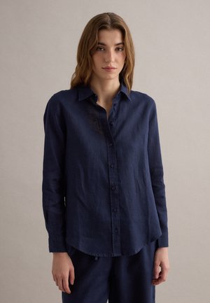 Jonge vrouw met lichtbruin golvend haar, gekleed in een marineblauw overhemd met knopen en bijpassende broek, staand tegen een effen beige achtergrond.