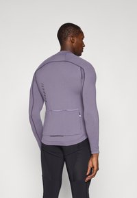 Camiseta deportiva de manga larga morada con un diseño ajustado, que cuenta con un bolsillo con cremallera en la parte posterior y detalles de costura en contraste.