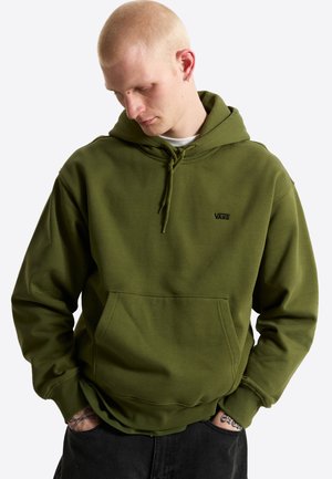 Giovane uomo con pelle chiara e capelli biondi corti che indossa una felpa con cappuccio Vans verde oliva, con le mani nelle tasche, guarda in basso a sinistra.