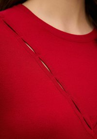 Czerwony sweter z dzianiny z ukośnymi wycięciami po jednej stronie, o gładkiej fakturze i dopasowanym kroju.
