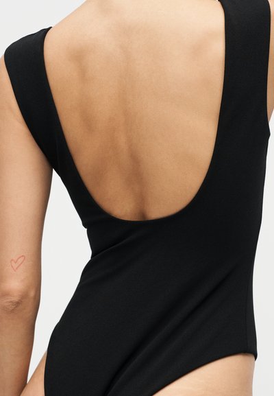 Γυναίκα που φοράει μαύρο, αμάνικο κορμάκι με βαθύ στρογγυλό άνοιγμα πίσω, που αφήνει εκτεθειμένο το πάνω μέρος της πλάτης και ένα μικρό κόκκινο τατουάζ καρδιάς στο αριστερό χέρι.