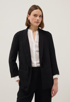 Frau mit kurzen braunen Haaren, die goldene Ohrringe trägt, ein strukturiertes schwarzes Blazer, weiße Bluse mit dezenten Punkten und schwarze Hose vor einfachem Hintergrund.