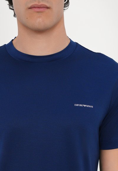 Tamsiai mėlynas trumpas rankovėmis marškinėliai pagaminti iš lygaus audinio. Sukurta su apvalia kaklo iškirpte ir mažuoju baltu "Emporio Armani" logotipu ant krūtinės.