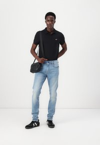 Moški nosi črno polo majico, svetlo modre kavbojke, črne superge in nosi črno torbico čez ramo na enotni beli podlagi.