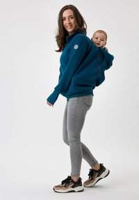 Felpa in pile turchese con tasca a marsupio e cerniera, con una sezione per trasportare il bambino sul retro. Indossata con jeans grigi e sneakers multicolori.