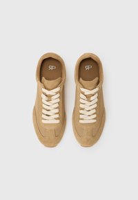Tan suede sneakers with beige laces, rounded toe, and subtle stitching details. The insole features a logo in white against a brown background.