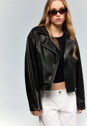 Mladá žena s dlhými blond vlasmi, ktorá nosí tmavé slnečné okuliare, čiernu koženú bundu, čierny crop top a biele nohavice na sivom pozadí.