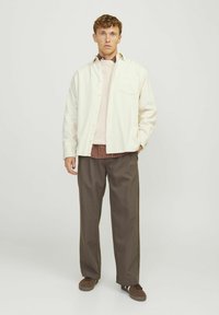 Camicia a bottoni di colore chiaro sopra un maglione rosa e una camicia a quadri, abbinata a pantaloni marroni e scarpe marroni. Sfondo neutro.