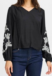 Blouse noire à manches longues avec décolleté en V. Présente des broderies florales blanches sur les manches. Ourlet droit, tissu texturé. Portée avec un jean foncé.