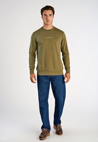 Oliwkowy bawełniany sweter z okrągłym dekoltem i logo Lindbergh, w połączeniu z niebieskimi dżinsami i brązowymi butami na sznurówki.