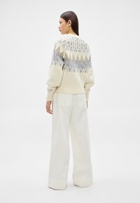 Mulher com cabelo liso e escuro, vestindo um suéter creme estampado e calças brancas de perna larga, em pé e de perfil sobre um fundo branco.