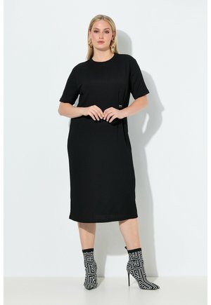 Kobieta stojąca na tle białej ściany, ubrana w czarną sukienkę midi, złote kolczyki koła oraz czarno-białe botki na wysokim obcasie w wzory.