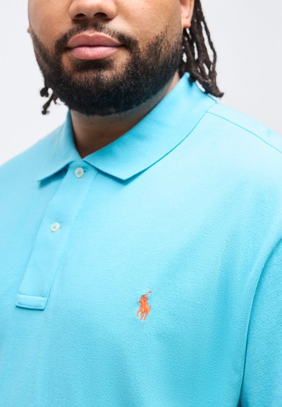 Άνδρας με γένια που φοράει ένα ανοιχτό μπλε πόλο μπλουζάκι με πορτοκαλί κεντημένο λογότυπο παίκτη πόλο στο στήθος, μπροστά σε απλό φόντο.