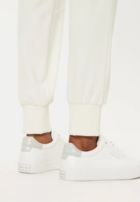 Sneakers bianchi con tomaia in pelle liscia, dettagli grigi nel tallone e suola in gomma testurizzata. Abbinati a joggers crema alle caviglie.