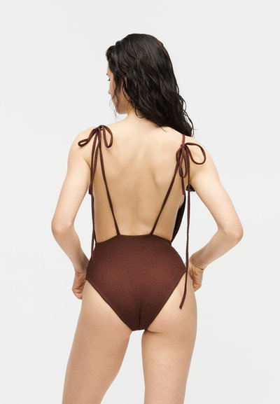 Femme aux longs cheveux foncés portant un maillot une pièce brun texturé avec de fines attaches aux épaules et un dos ouvert, debout les mains sur les hanches.