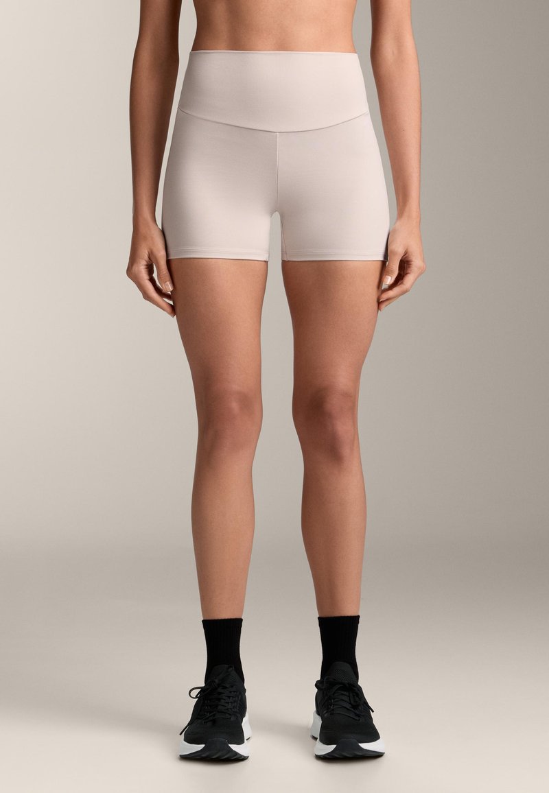 Svetlo béžové šortky s vysokým pásom, hladkou textúrou, priliehajúcim strihom a dĺžkou do polovice stehien. Nosené s čiernymi teniskami a ponožkami.