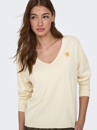 Jeune femme aux longs cheveux bruns portant un pull crème à col en V avec un emblème de soleil jaune sur la poitrine, debout devant un arrière-plan uni.