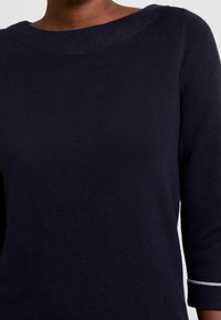 Pull bleu marine en tissu doux avec un large col, des manches trois-quarts et des coutures blanches le long des poignets. Texture lisse.