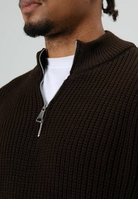 Bruine gebreide trui met een hoge kraag en een halve rits. Gestructureerde stof met zichtbare ribbels en zilverkleurige ritshardware.