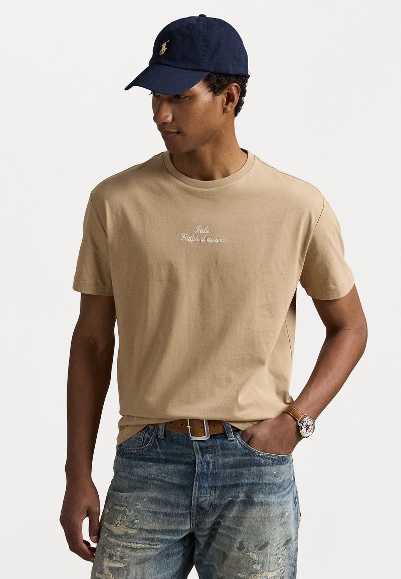 Bežové bavlnené tričko s vyšitým logom „Polo Ralph Lauren“, v kombinácii s poškodenými modrými džínami a námorníckou čiapkou.