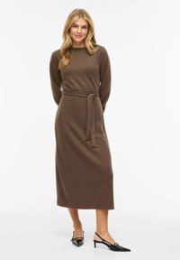Brązowa prążkowana sukienka dzianinowa z długimi rękawami i paskiem do wiązania. Sukienka ma długość midi, zestawiona z czarnymi czółenkami z ostrym noskiem.