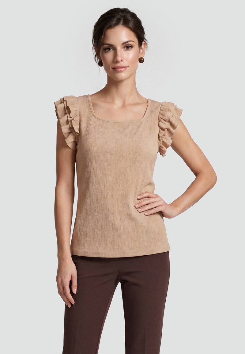 Femme aux cheveux foncés portant un haut beige à manches volantées et un pantalon brun foncé, debout avec une main sur la hanche devant un fond gris.