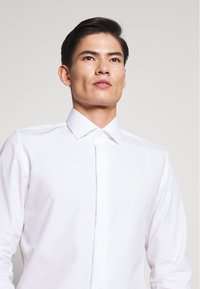Witte blouse met lange mouwen, een klassieke kraag en een gladde, matte textuur. Eenvoudig ontwerp zonder patronen of accenten.
