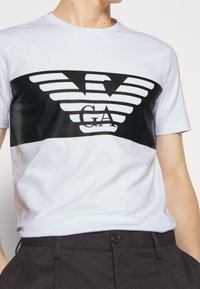 Man wearing white T-shirt with black horizontal logo featuring stylized eagle and "GA" letters, paired with dark trousers.
