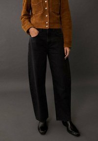 Ruskea mokkajakku helmi-napeilla, yhdistettynä korkeavyötäröisiin mustiin leveälahkeisiin farkkuihin ja mustiin kärkikengiin.