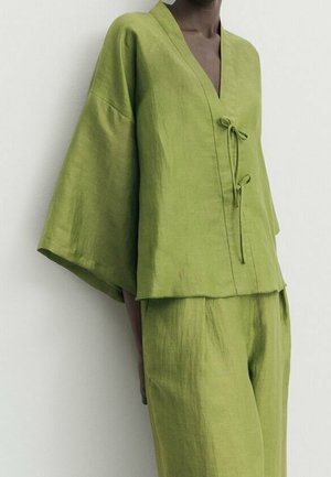 Model wearing loose-fitting green linen top with wide sleeves and matching high-waisted pants, standing against white background.