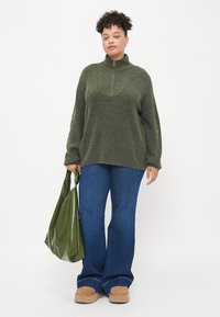 Suéter texturizado de color verde oliva con media cremallera, combinado con pantalones vaqueros de campana en azul oscuro. Sosteniendo un bolso tote brillante de color verde oscuro. Zapatos beige.