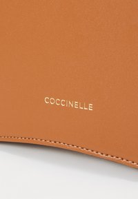 Tamna kožna torba sa urezanim logotipom "Coccinelle" u zlatnoj boji. Glatka tekstura s urednim šavovima i čistim ivicama.
