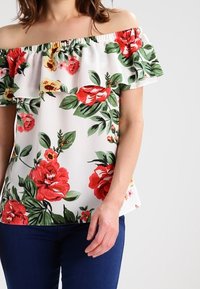 Kukkakuvioinen olkaimeton toppi, jossa on valkoinen tausta ja punaisia sekä vihreitä kukkia, röyhelökaula-aukko ja rento istuvuus. Yhdistetty tummansinisiin housuihin.