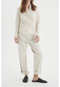 Kvinna klädd i en krämfärgad omslagströja med långa ärmar, ljusbeige uppvikta byxor och svarta platta skor utan snörning, stående mot vit bakgrund.