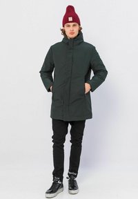 Donkergroene gewatteerde parka met een hoge kraag, zijzakken, gedragen over zwarte slim-fit broek en zwarte sneakers. Bevat een geribbelde bordeauxrode muts.