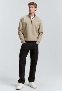 Béžová mikina na zips so stojacím golierom, kombinovaná s čiernymi džínsami a bielymi teniskami. Jednoduchý dizajn s dlhými rukávmi.