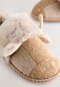 Chaussons beiges duveteux avec accents d'oreilles, mélange de textures de tissu doux et de motifs chevrons, ornés de points dorés et de bords cousus.