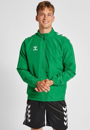 Groene lichtgewicht jas met een volledige ritssluiting aan de voorkant, elastische manchetten en witte accenten op de schouders. Gepaard met zwarte korte broek met witte chevron.