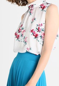 Blouse sans manches florale blanche avec des motifs roses et verts, présentant un col haut et un matériau doux, associée à une jupe plissée bleue.