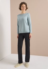 Light blue, long-sleeve top with a round neck, paired with black pants and light sneakers. Slim fit with gathered design at the waist.