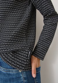 Top a maniche lunghe con motivo bianco e nero e tessuto testurizzato, dotato di un piccolo etichetta del marchio. Indossato con jeans in denim blu.