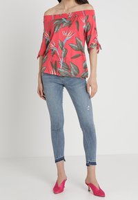 Top rojo con escote ligeramente caído, estampado floral y detalles de lazo, combinado con unos vaqueros desgastados de color azul claro y mules rosas de tacón.