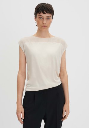 Top corto a maniche corte di un beige chiaro con sovrapposizione trasparente sulle spalle, texture liscia e vestibilità comoda. Indossato con pantaloni neri su misura.
