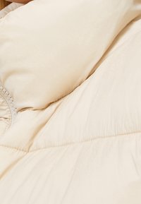 Cream-colored puffer jacket with a smooth texture, featuring quilted sections and subtle stitching details along the seams.